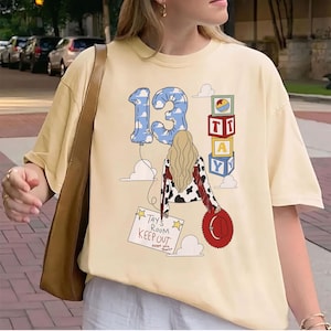 K&ouml;nnte beinhalten: Ein cremefarbenes T-Shirt mit einer Grafik einer Person mit langem blondem Haar, einer Kuhfell-Jacke und einem roten Hut. Das Design zeigt einen blauen, wolkenmusterartigen Luftballon mit der Zahl 13, Spielw&uuml;rfel und ein Schild TAY'S ROOM KEEP OUT.