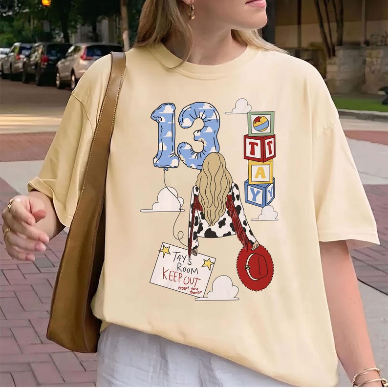 K&ouml;nnte beinhalten: Ein cremefarbenes T-Shirt mit einer Grafik einer Person mit langem blondem Haar, einer Kuhfell-Jacke und einem roten Hut. Das Design zeigt einen blauen, wolkenmusterartigen Luftballon mit der Zahl 13, Spielw&uuml;rfel und ein Schild TAY'S ROOM KEEP OUT.