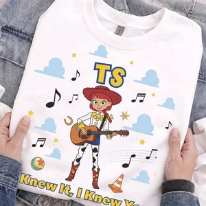Peut inclure: Un t-shirt blanc &agrave; col rond orn&eacute; d'un personnage de dessin anim&eacute; portant un chapeau rouge et un pantalon &agrave; motifs de vache, jouant de la guitare acoustique. Le motif inclut des nuages bleus, des &eacute;toiles jaunes, des notes de musique et du texte TS et Knew It, I Knew You.