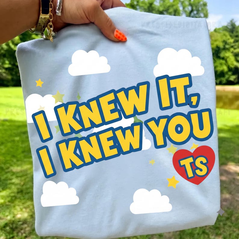Puede incluir: Una camiseta azul claro con un estampado gr&aacute;fico de nubes blancas y estrellas amarillas. El texto "I KNEW IT, I KNEW YOU" aparece en letras amarillas en negrita con un borde azul oscuro. Un coraz&oacute;n rojo contiene las letras amarillas "TS".