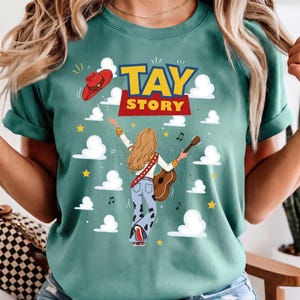 Puede incluir: Una camiseta color verde azulado con el gr&aacute;fico de una persona de cabello rubio largo tocando una guitarra ac&uacute;stica. El dise&ntilde;o incluye un sombrero de vaquero rojo, nubes blancas, estrellas amarillas, notas musicales y el texto TAY STORY.