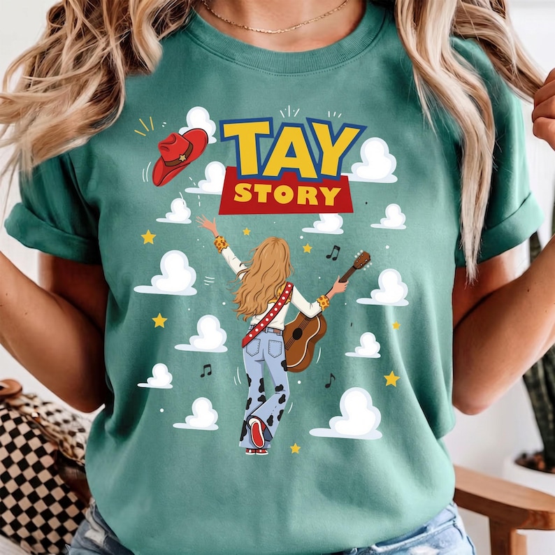 Puede incluir: Una camiseta color verde azulado con el gr&aacute;fico de una persona de cabello rubio largo tocando una guitarra ac&uacute;stica. El dise&ntilde;o incluye un sombrero de vaquero rojo, nubes blancas, estrellas amarillas, notas musicales y el texto TAY STORY.