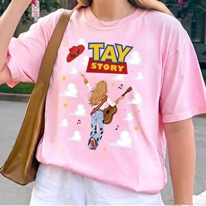 Puede incluir: Una camiseta rosa claro con un estampado gr&aacute;fico de una persona con cabello largo y rubio tocando una guitarra ac&uacute;stica, vistiendo pantalones acampanados con estampado de vaca. Arriba hay un sombrero de vaquero rojo y el texto TAY STORY.
