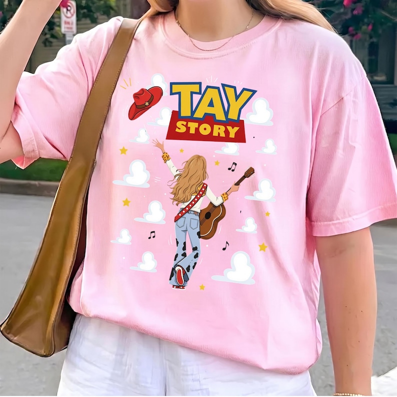 Puede incluir: Una camiseta rosa claro con un estampado gr&aacute;fico de una persona con cabello largo y rubio tocando una guitarra ac&uacute;stica, vistiendo pantalones acampanados con estampado de vaca. Arriba hay un sombrero de vaquero rojo y el texto TAY STORY.