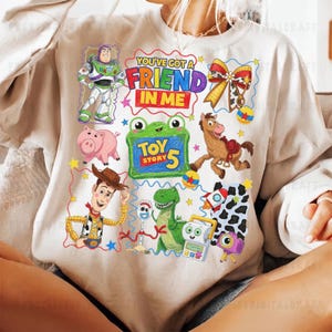 May include: A beige crewneck sweatshirt featuring a colorful graphic print of Toy Story characters including Buzz Lightyear, Woody, Rex, and Hamm. The design includes the text "YOU'VE GOT A FRIEND IN ME" and "TOY STORY 5" surrounded by stars, a bow, and various small icons.