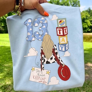 K&ouml;nnte beinhalten: Ein hellblaues T-Shirt mit einer Grafik einer Person mit blondem Haar, einer Kuhfell-Jacke mit roten Fransen und einem roten Hut. Das Design zeigt einen Ballon in Form der Zahl 13, Buchstabenw&uuml;rfel und ein Schild mit der Aufschrift TAY'S ROOM KEEP OUT.