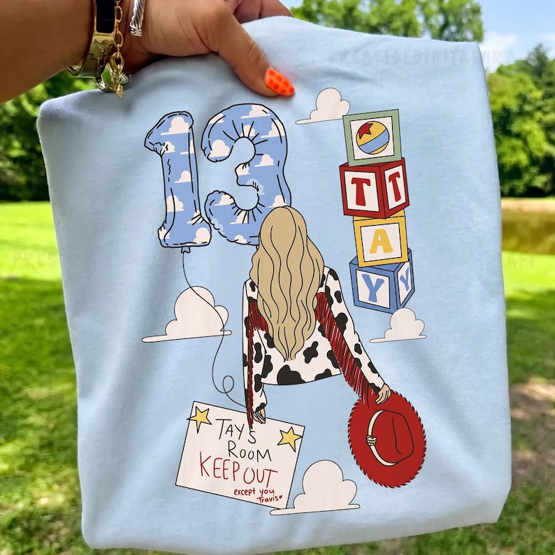 K&ouml;nnte beinhalten: Ein hellblaues T-Shirt mit einer Grafik einer Person mit blondem Haar, einer Kuhfell-Jacke mit roten Fransen und einem roten Hut. Das Design zeigt einen Ballon in Form der Zahl 13, Buchstabenw&uuml;rfel und ein Schild mit der Aufschrift TAY'S ROOM KEEP OUT.
