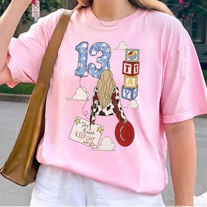 K&ouml;nnte beinhalten: Ein hellrosa T-Shirt mit einer Grafik einer Person mit blondem Haar, einer Jacke mit Kuhmuster und einem roten Hut. Das Design zeigt einen blauen, wolkenbedruckten Luftballon in Form einer 13, Spielw&uuml;rfel und ein Schild mit Tay's Room KEEP OUT.