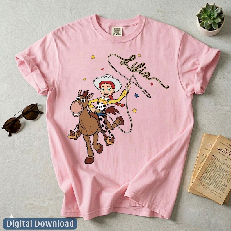 May include: A light pink t-shirt featuring a graphic of a cowgirl character riding a brown horse. The character wears a white hat, yellow shirt, and cow-print vest. The name Lilia is written in cursive script above the character with a lasso rope.