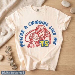 Op de afbeelding: Een cr&egrave;mekleurig T-shirt met korte mouwen en een rode lijntekening van twee omhelzende figuren, met de blauwe tekst "YOU'RE A COWGIRL LIKE ME" in een boog erboven en de gele en blauwe letters "TS" onder de figuren geplaatst.