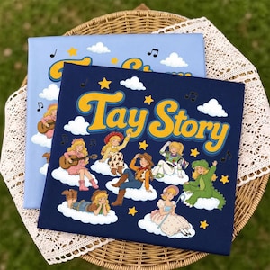 May include: A dark blue rectangular canvas print featuring the text Tay Story in yellow with a blue outline. The design includes multiple illustrations of a blonde character dressed in various costumes sitting on white clouds with yellow stars.