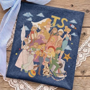 May include: A dark blue heathered t-shirt featuring a graphic print of multiple illustrated characters in various outfits, including a space ranger suit and cowgirl attire, with the letters TS, a rocket ship, a ball, and a sheriff star badge.