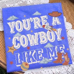 May include: A blue t-shirt featuring the text "YOU'RE A COWBOY LIKE ME" in white letters. The design includes a brown cowboy hat, a rocket, a sheriff star badge, a ball, and brown cowboy boots, accented with small stars and light blue clouds.