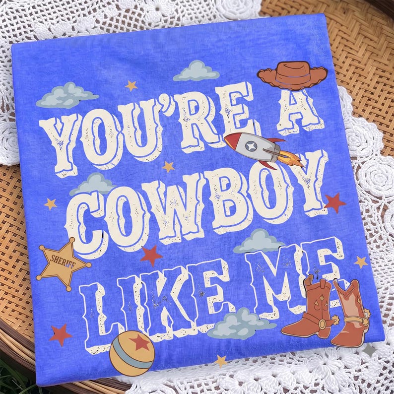 May include: A blue t-shirt featuring the text "YOU'RE A COWBOY LIKE ME" in white letters. The design includes a brown cowboy hat, a rocket, a sheriff star badge, a ball, and brown cowboy boots, accented with small stars and light blue clouds.