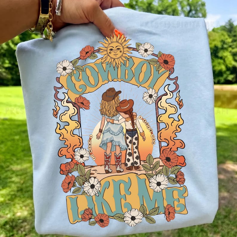 May include: A light blue t-shirt featuring a graphic print of two figures in cowboy hats standing together. The design includes a sun, flowers, and the text "COWBOY LIKE ME" and "I KNEW YOU". The figures wear western-style clothing and boots.