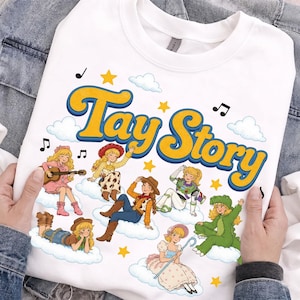 May include: A white crewneck t-shirt featuring the text Tay Story in yellow with blue outlines. The graphic includes multiple illustrations of a blonde character dressed in various costumes, including a cowgirl, space ranger, and dinosaur, on clouds.