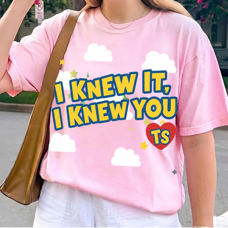 Puede incluir: Una camiseta rosa claro con el texto amarillo I KNEW IT, I KNEW YOU con un borde azul. El dise&ntilde;o incluye nubes blancas, peque&ntilde;as estrellas amarillas y un coraz&oacute;n rojo con las letras TS en amarillo. La camiseta tiene mangas cortas.