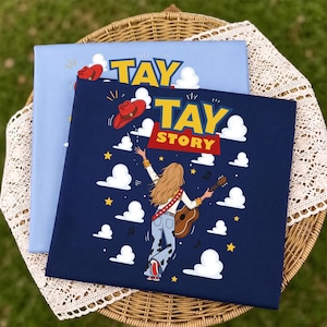 Puede incluir: Una camiseta azul marino oscuro con un gr&aacute;fico de una persona con cabello largo rubio tocando una guitarra ac&uacute;stica, usando un sombrero de vaquero rojo y jeans azul claro. El texto TAY TAY STORY est&aacute; impreso arriba, rodeado de nubes blancas y estrellas amarillas.