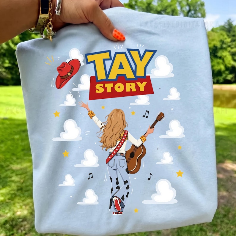 Puede incluir: Una camiseta azul claro con el gr&aacute;fico de una persona de pelo rubio largo tocando una guitarra ac&uacute;stica. El dise&ntilde;o incluye el texto TAY STORY, un sombrero de vaquero rojo, nubes blancas, estrellas amarillas y notas musicales sobre fondo azul.