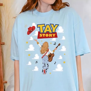 Puede incluir: Una camiseta azul claro con un gr&aacute;fico de una persona con cabello largo y rubio vista desde atr&aacute;s, vistiendo una blusa blanca y pantalones con estampado de vaca, sosteniendo una guitarra. El texto TAY STORY est&aacute; impreso arriba, con un sombrero vaquero.