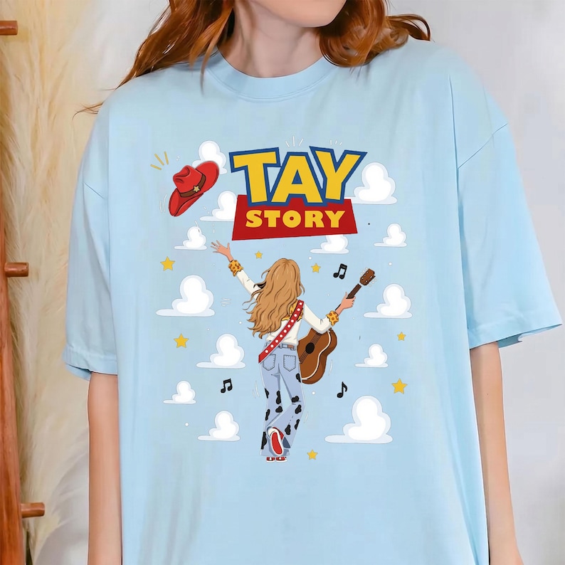 Puede incluir: Una camiseta azul claro con un gr&aacute;fico de una persona con cabello largo y rubio vista desde atr&aacute;s, vistiendo una blusa blanca y pantalones con estampado de vaca, sosteniendo una guitarra. El texto TAY STORY est&aacute; impreso arriba, con un sombrero vaquero.