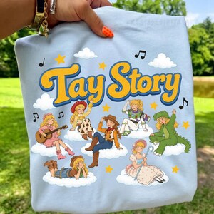 May include: A light blue t-shirt featuring the yellow text Tay Story with a dark blue outline. The graphic includes seven cartoon characters dressed in various costumes, sitting on white clouds, surrounded by yellow stars and black musical notes.