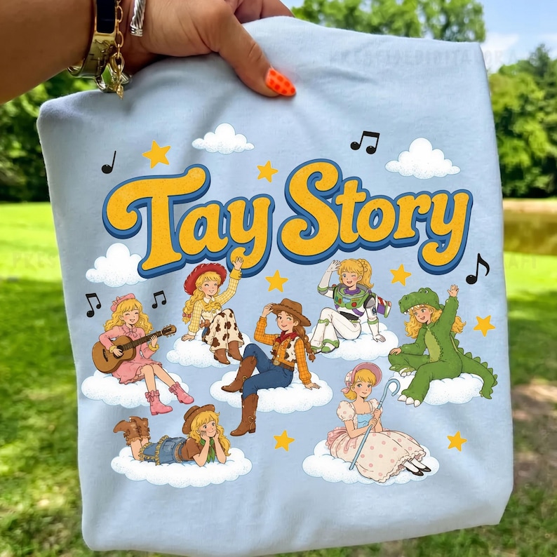 May include: A light blue t-shirt featuring the yellow text Tay Story with a dark blue outline. The graphic includes seven cartoon characters dressed in various costumes, sitting on white clouds, surrounded by yellow stars and black musical notes.