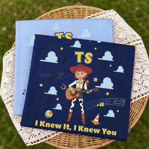 Peut inclure: Un panneau en tissu carr&eacute; bleu fonc&eacute; repr&eacute;sentant un personnage de dessin anim&eacute; aux cheveux roux jouant de la guitare acoustique, entour&eacute; de nuages blancs, d'&eacute;toiles jaunes, de notes de musique, d'un ballon et d'un c&ocirc;ne, avec le texte TS et I KNEW IT, I KNEW YOU.