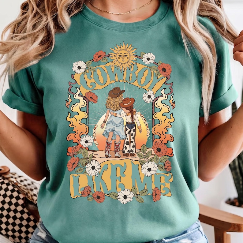 May include: A teal short-sleeve t-shirt featuring a graphic print of two figures in cowboy hats with arms around each other. The design includes a sun, flowers, and the text "COWBOY LIKE ME" in a bold, yellow, retro-style font on the front.