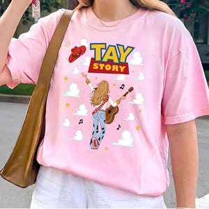 Puede incluir: Una camiseta rosa claro con un estampado gr&aacute;fico que incluye el texto TAY STORY en amarillo y rojo. El dise&ntilde;o presenta un sombrero de vaquero rojo, nubes blancas, estrellas amarillas, notas musicales y una ilustraci&oacute;n tocando la guitarra.