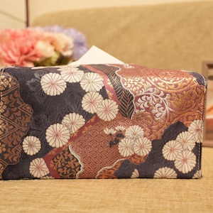 Può includere: Un copri scatola di fazzoletti decorativo con un motivo floreale e geometrico. Il copri scatola presenta uno sfondo blu scuro con fiori bianchi e rossi e accenti dorati e marroni. La scatola è rettangolare.