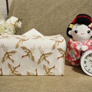 Può includere: Un portatovaglioli bianco con un motivo di gru dorate, una bambola di peluche in kimono e un orologio bianco. L'orologio ha numeri e lancette nere. Su una superficie beige.