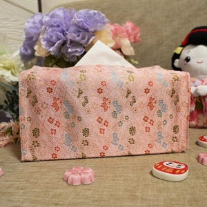 Può includere: Un copri scatola di fazzoletti rosa con un motivo floreale, fazzoletti bianchi e una bambola geisha di peluche. Saponi decorativi rosa a forma di fiore e una bambola Daruma in primo piano. Fiori viola e rosa sullo sfondo.