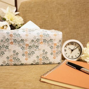 Può includere: Un copri scatola di fazzoletti rettangolare con un motivo floreale bianco, grigio e arancione. Un orologio analogico bianco, una penna e un taccuino a spirale sono accanto alla scatola di fazzoletti. Fiori decorativi sullo sfondo.