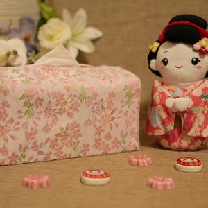 Può includere: Un copri scatola di fazzoletti rosa floreale e una bambola in kimono. La bambola ha uno chignon nero e indossa un kimono rosso e rosa. Davanti ci sono diverse caramelle rosa a forma di fiore e una piccola caramella rotonda.