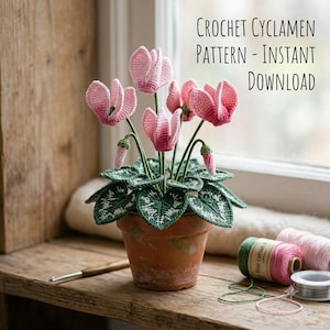 May include: A crochet cyclamen plant in a terracotta pot with pink flowers and green leaves featuring white veins. The text reads: CROCHET CYCLAMEN PATTERN - INSTANT DOWNLOAD. A crochet hook and two spools of yarn are visible nearby.