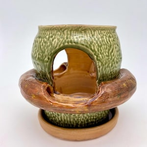 Peut inclure: Un brûle-parfum en céramique composé d'une partie supérieure verte texturée à la finition brillante et d'une base annulaire marron lisse à l'aspect bois. La structure présente une ouverture centrale et repose sur un plateau circulaire marron.