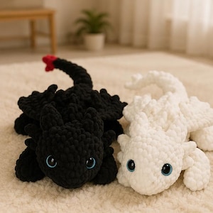 May include: Two plush dragon toys made of chunky yarn. One dragon is solid black with a small red tip on its tail, and the other is solid white. Both creatures feature large, round, light blue eyes and textured, soft, crocheted bodies.