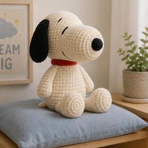 May include: A cream-coloured crochet plush toy shaped like a dog with a large black ear, a black nose, and a red collar. The figure has a rounded head, a prominent snout, and a seated posture with visible stitched texture throughout the body.