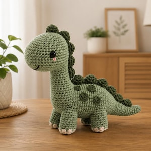 May include: A sage green crochet dinosaur plush toy featuring a long neck, a row of dark green textured spikes along its back, round dark green spots on its side, black eyes, and small light beige accents on the feet and a pink blush detail.