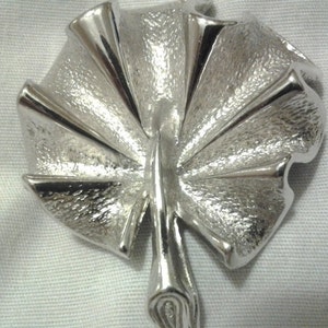 May include: A silver-toned brooch in the shape of a stylized leaf. The brooch has a textured surface and a fan-like design with multiple segments radiating from a central stem. The metal has a bright, reflective quality.