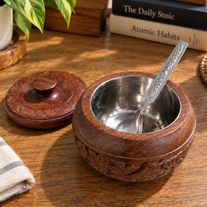 Puede incluir: Un recipiente redondo de madera con acabado marrón oscuro y tallas florales intrincadas en el exterior. Cuenta con una tapa extraíble, un cuenco interior de acero inoxidable pulido y una cuchara de metal plateado a juego con motivos de hojas.