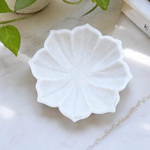 May include: A white marble bowl carved into the shape of a blooming lotus flower with eight distinct petals. The bowl features a smooth, matte finish and a shallow, concave center, showcasing the natural crystalline texture of the stone material.