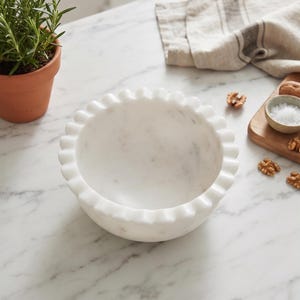 May include: A white marble bowl featuring a scalloped, ruffled edge design. The bowl has a smooth, polished interior and exterior surface with natural grey veining throughout the stone, creating a distinct pattern on the circular vessel.