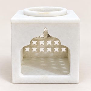 May include: A white marble candle holder shaped as a cube with a circular opening on top. The front features an arched cutout with an intricate lattice pattern of small cross-shaped perforations, allowing light to pass through the stone structure.
