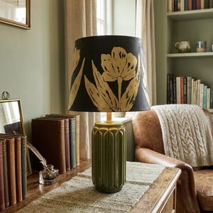 Può includere: Lampada da tavolo in ceramica verde con paralume nero con design a tulipani dorati. La lampada è su un tavolo di legno con libri e un runner decorativo. Atmosfera vintage.
