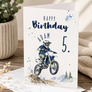 Könnte beinhalten: Eine weiße Geburtstagskarte mit einer Aquarellillustration einer Person auf einem blau-weißen Motocross-Motorrad. Der Text lautet Happy Birthday, Adam, und die Zahl 5. Das Design enthält blaue und braune Farbspritzer.