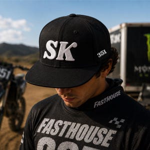 May include: Black baseball cap with the white letters "SK" on the front. The number "351" is on the side of the cap. The person in the image is wearing a black shirt with the word "Fasthouse" printed on it.