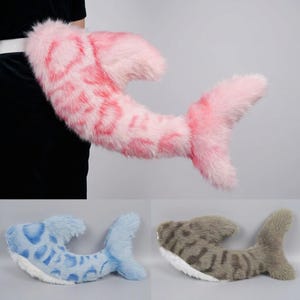 May include: A plush faux fur tail accessory shaped like a shark, featuring a pink and white spotted pattern, a blue and white spotted version, and a brown and white striped version, each with a prominent dorsal fin and a wide, forked tail fin.