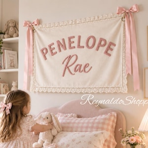 May include: A rectangular cream-colored fabric wall hanging featuring the name Penelope Rae in raised dusty pink letters. The banner is trimmed with white lace and suspended by two dusty pink satin ribbons tied in bows at the top corners.